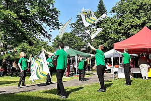 Gruppe von Menschen in grünen T-Shirts wirft Fahnen in die Luft auf einer Wiese mit Zelten und Bäumen im Hintergrund