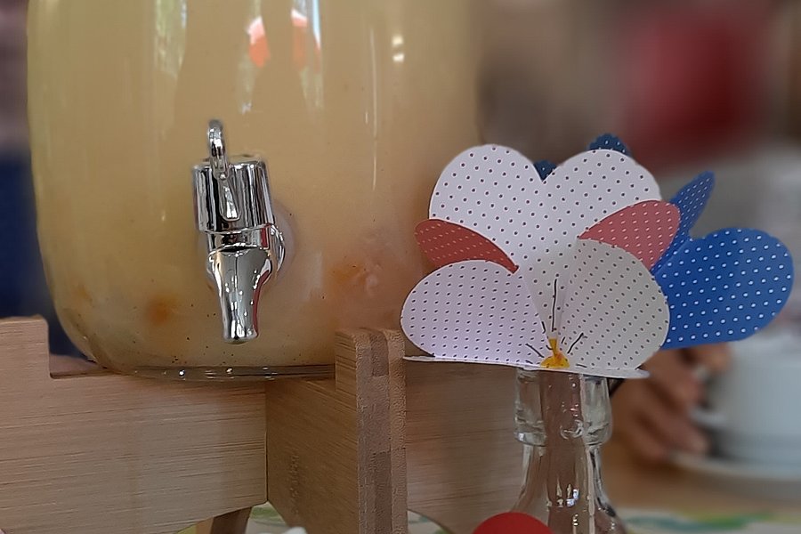 Glasbehälter mit Zapfhahn gefüllt mit hellem Getränk und Glas mit Papierherzen daneben im unscharfen Hintergrund
