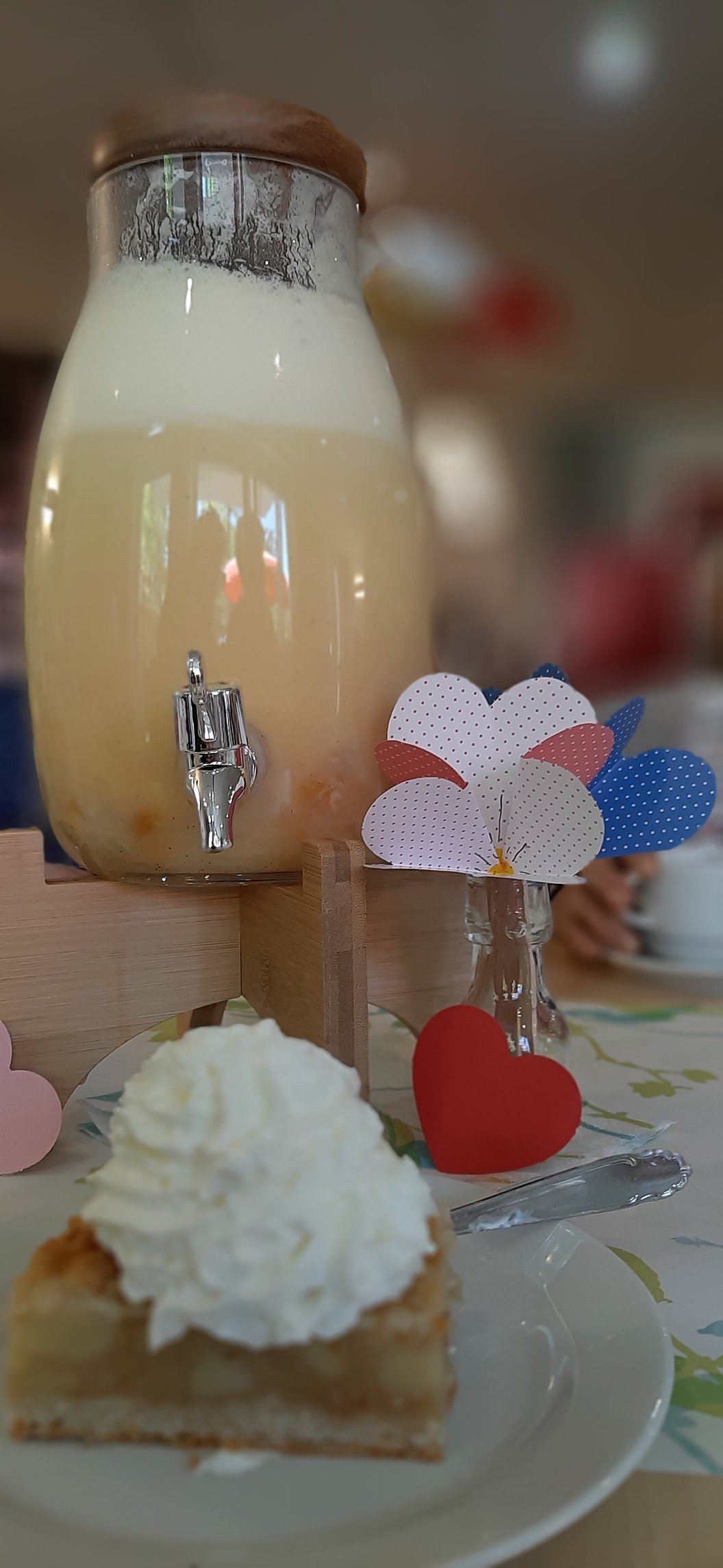 Glasbehälter mit Zapfhahn gefüllt mit hellem Getränk und Glas mit Papierherzen daneben im unscharfen Hintergrund