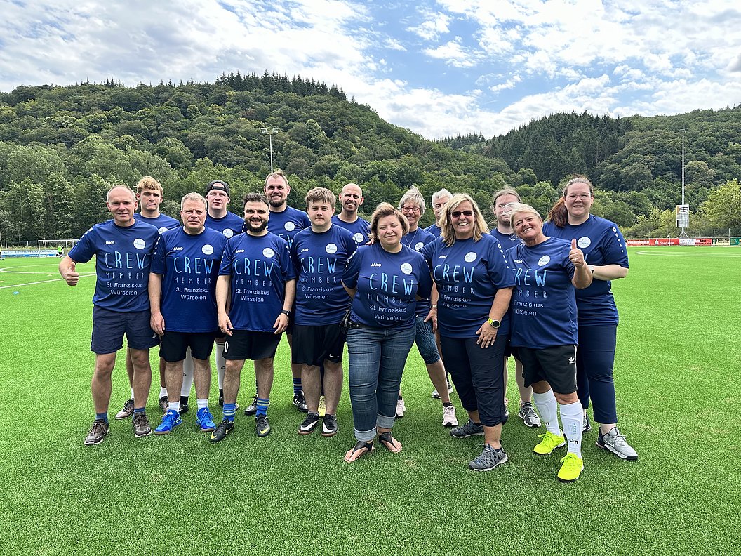 Gruppe von Menschen in blauen T-Shirts mit der Aufschrift 'CREW' steht auf einem Sportplatz vor bewaldeten Hügeln