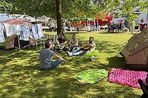 Gruppe von vier Personen sitzt auf einer Wiese unter Bäumen, umgeben von Zelten und Wäscheleinen mit aufgehängter Kleidung