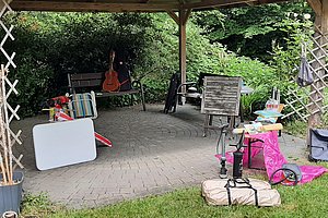 Überdachte Terrasse mit Gartenmöbeln, einem weißen Brett, einer Gitarre und verstreuten Gegenständen auf gepflastertem Boden und Rasen