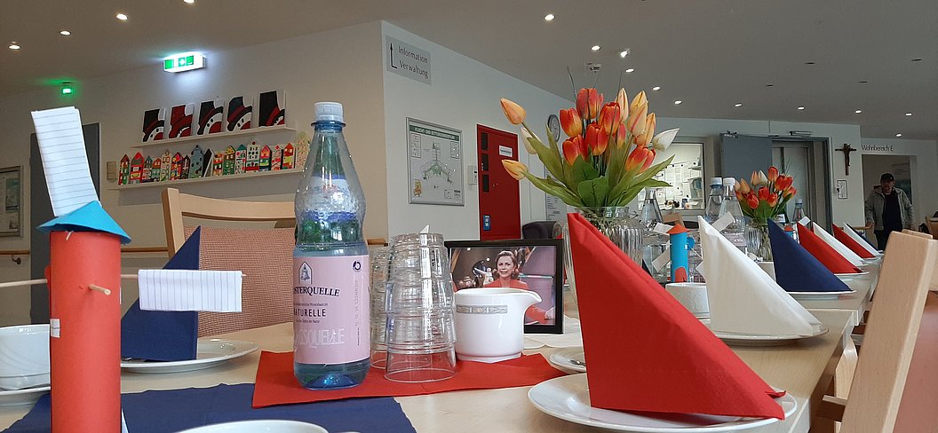 Tisch mit rot gefalteten Servietten, Wasserflasche, Plastikbechern und Blumenstrauß in einem hellen Raum mit Stühlen und Bildern an der Wand