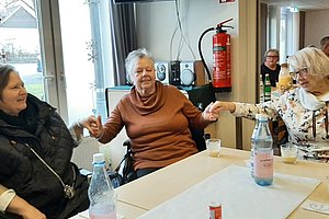 Drei ältere Frauen sitzen an einem Tisch und halten sich an den Händen, auf dem Tisch stehen Wasserflaschen und Gläser.