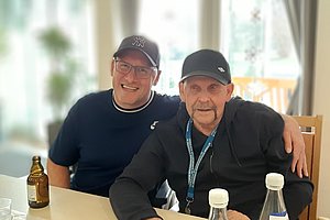 Zwei Männer sitzen an einem Tisch, lächeln und genießen Getränke in einer hellen Umgebung.