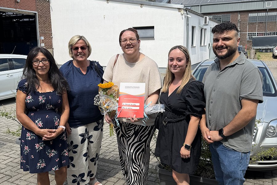 Fünf Personen stehen vor einem weißen Gebäude auf gepflastertem Boden, eine Person hält eine rote Urkunde und einen Blumenstrauß