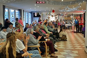 Gruppe älterer Menschen sitzt in einem Raum mit Kachelfußboden und großen Fenstern und schaut auf eine Person, die vorne spricht oder singt