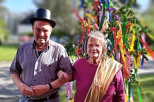 Zwei Personen gehen Arm in Arm auf einem Weg, eine trägt einen schwarzen Hut und kariertes Hemd, die andere eine goldfarbene Schärpe und ein Diadem