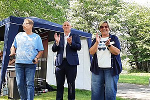 Drei Personen stehen im Freien vor einem blauen Pavillon, zwei klatschen, eine hält ein Mikrofon, im Hintergrund weitere Menschen und ein Rollstuhlfahrer auf einem Weg im Park