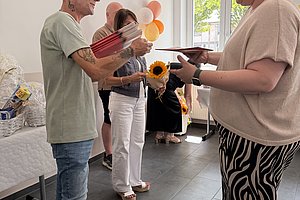 Drei Personen stehen in einem hellen Raum, eine Person überreicht einer anderen ein Geschenk, im Hintergrund sind Luftballons und eine Sonnenblume sichtbar