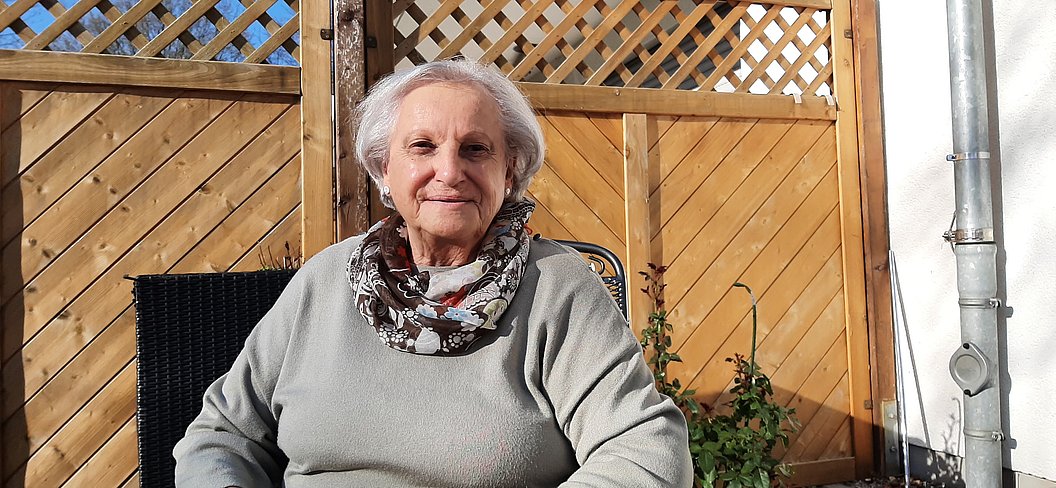Ältere Dame lächelt, sitzt auf einer Terrasse mit Holzlattenzaun, trägt einen grauen Pullover und einen Schal.