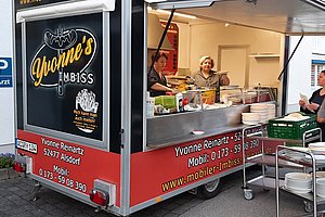 Imbisswagen mit der Aufschrift 'Yvonne's Imbiss' und zwei Personen hinter dem Verkaufstresen sowie zwei Personen vor dem Wagen mit Geschirr und Tabletts