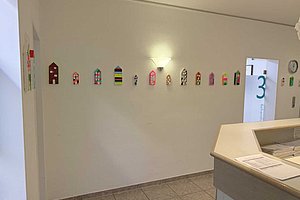 Flur mit einer Reihe bunter, kleiner Hausformen an der Wand über einer hellen Fliesenbodenfläche und einem Empfangstresen im Vordergrund