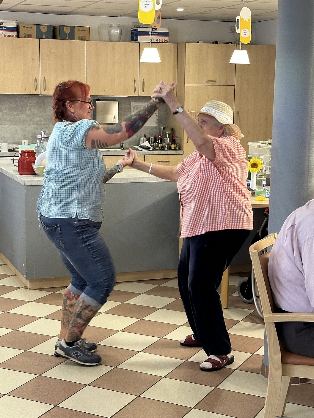 Zwei Frauen tanzen in einer Küche, eine trägt ein blaues Hemd und Jeans mit Tattoos, die andere ein rosa Oberteil und einen Hut.