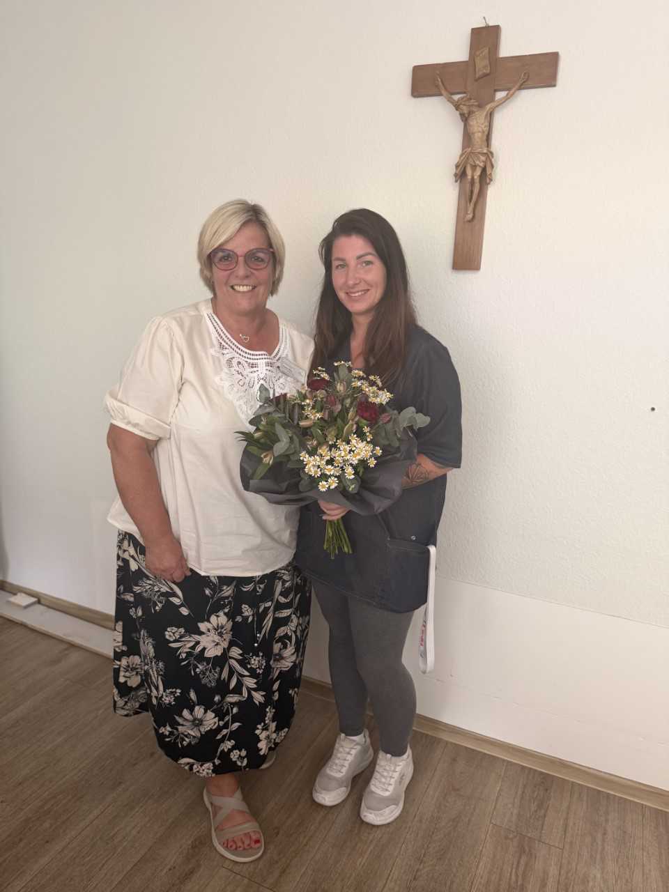 Zwei Frauen stehen vor einer weißen Wand mit einem Kruzifix, die jüngere hält einen Blumenstrauß